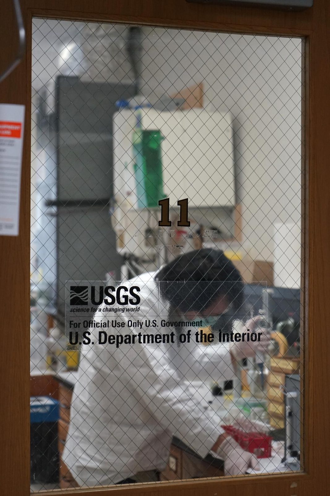 A research laboratory seen from outside through a glass window, with a scientist inside preparing samples for analysis. Real USGS photograph used on the Ellis Abel Learns "Why I Love Science" page.